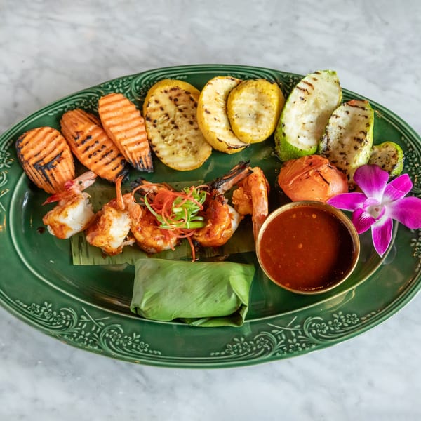 Grilled shrimp, carrots, zucchini, and tomatoes with dipping sauce and a purple orchid on a green plate.