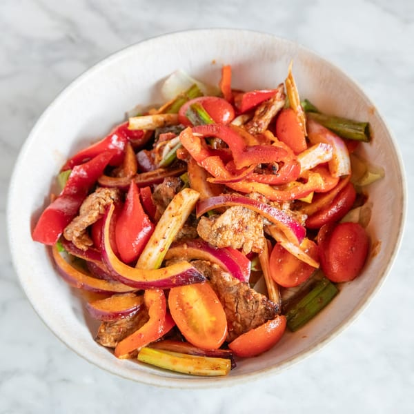 Close-up of a vibrant Thai stir-fry with beef, red peppers, onions, and tomatoes in a white bowl.