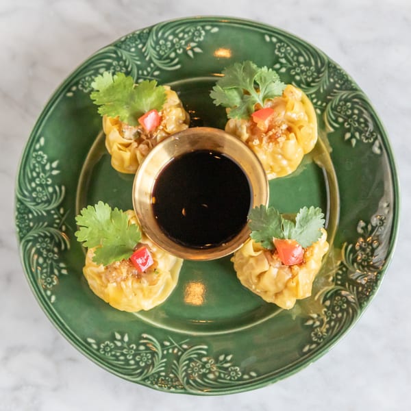 Four Thai dumplings with cilantro and red pepper on a green plate with a gold bowl of sauce.